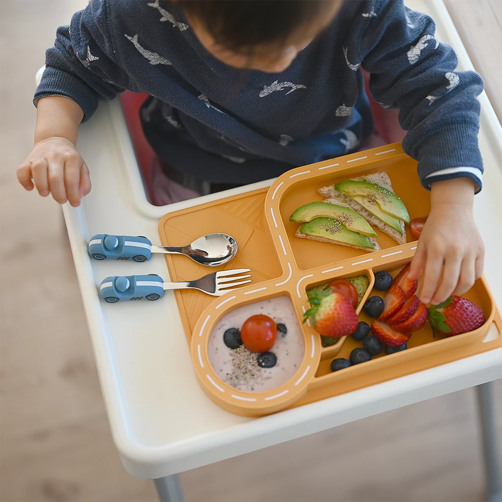 A toddler enjoying a meal with a yellow silicone car-themed dinnerware set featuring a racetrack plate design, complete with avocado toast, strawberries, blueberries, and yogurt. Accompanied by a playful blue car-shaped spoon and fork, this dinnerware makes mealtime fun and engaging for kids.