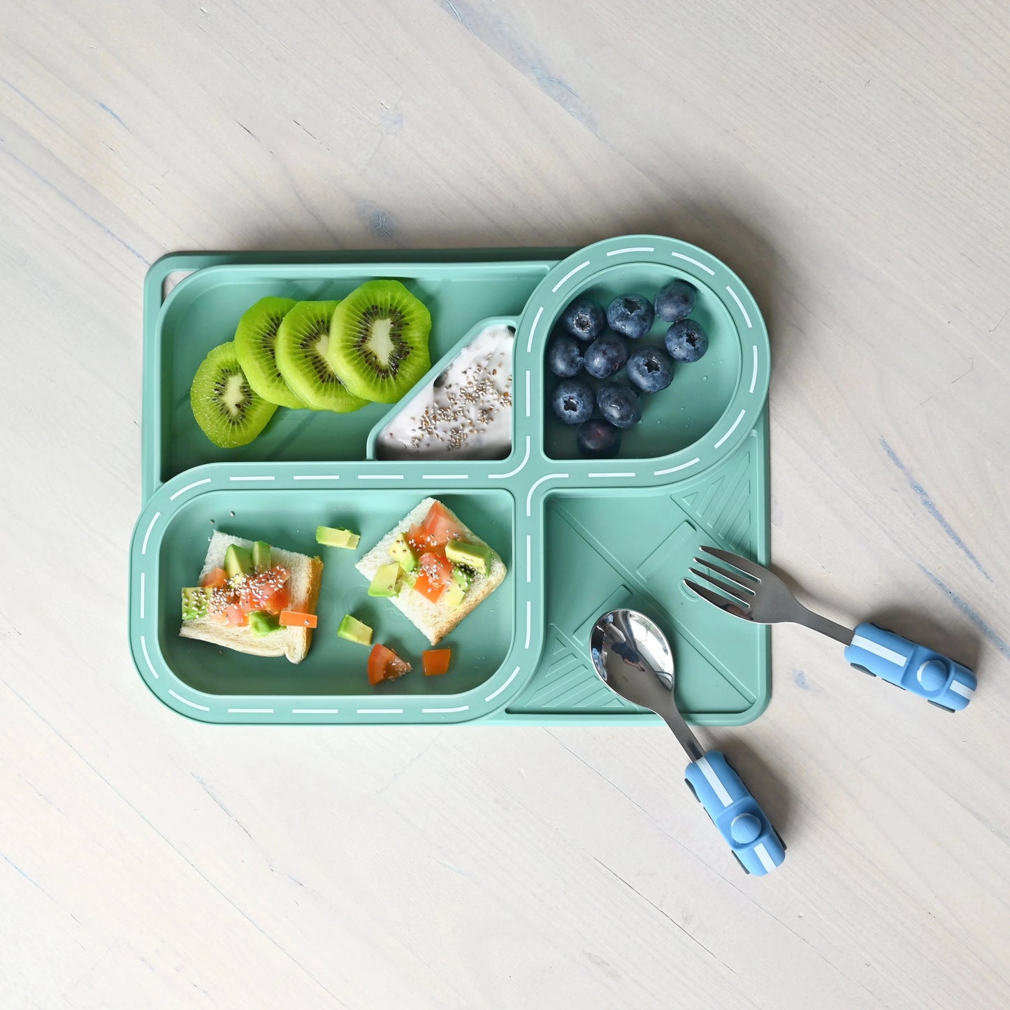 Children's segmented plate containing food, a fork, and spoon on a light wooden surface.