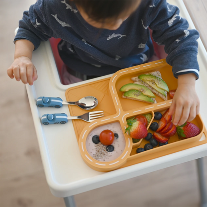 A toddler enjoying a meal with a yellow silicone car-themed dinnerware set featuring a racetrack plate design, complete with avocado toast, strawberries, blueberries, and yogurt. Accompanied by a playful blue car-shaped spoon and fork, this dinnerware makes mealtime fun and engaging for kids.