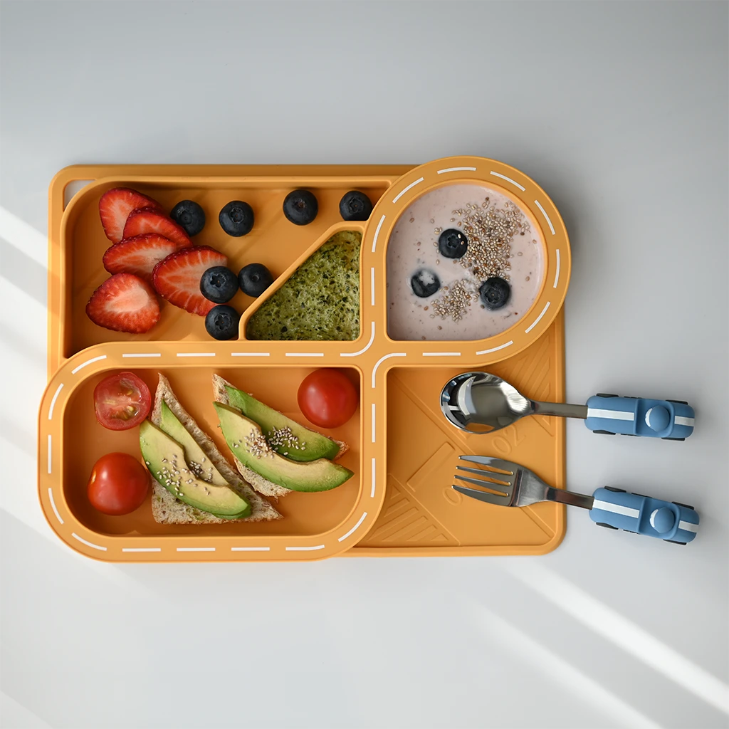 A beautifully arranged meal on a yellow silicone racetrack plate for kids, featuring fresh fruits like strawberries and blueberries, avocado toast, and yogurt. The car-themed dinnerware, with its playful blue spoon and fork, encourages fun and creativity during meals.